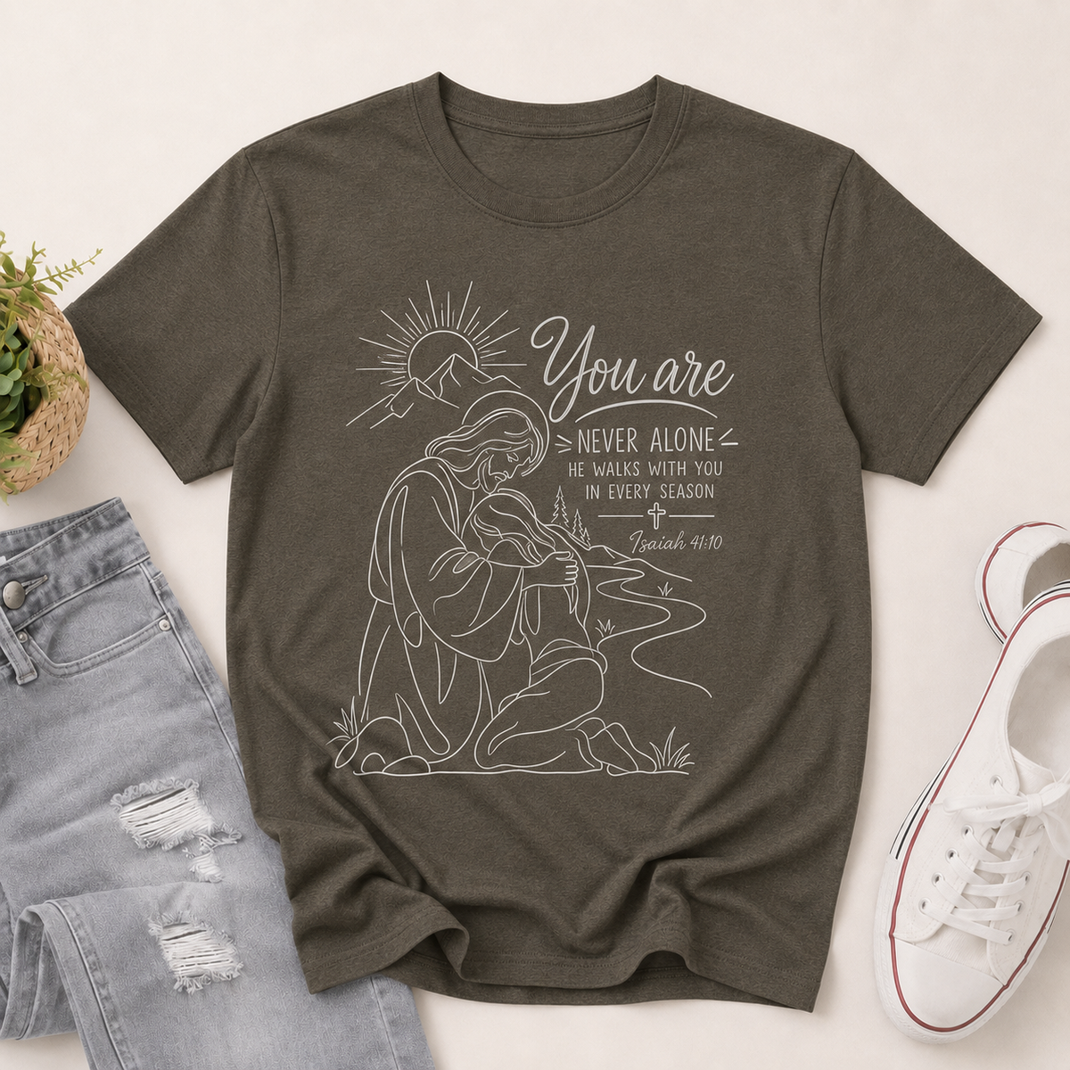 A dark olive green graphic t-shirt laid flat on a light beige background, styled with light-wash distressed denim jeans, a small potted succulent, and a white low-top sneaker. The t-shirt features a white line art graphic of a kneeling woman with a sunburst and the text 'You are NEVER ALONE HE WALKS WITH YOU IN EVERY Season Psalm 46:1-10'.