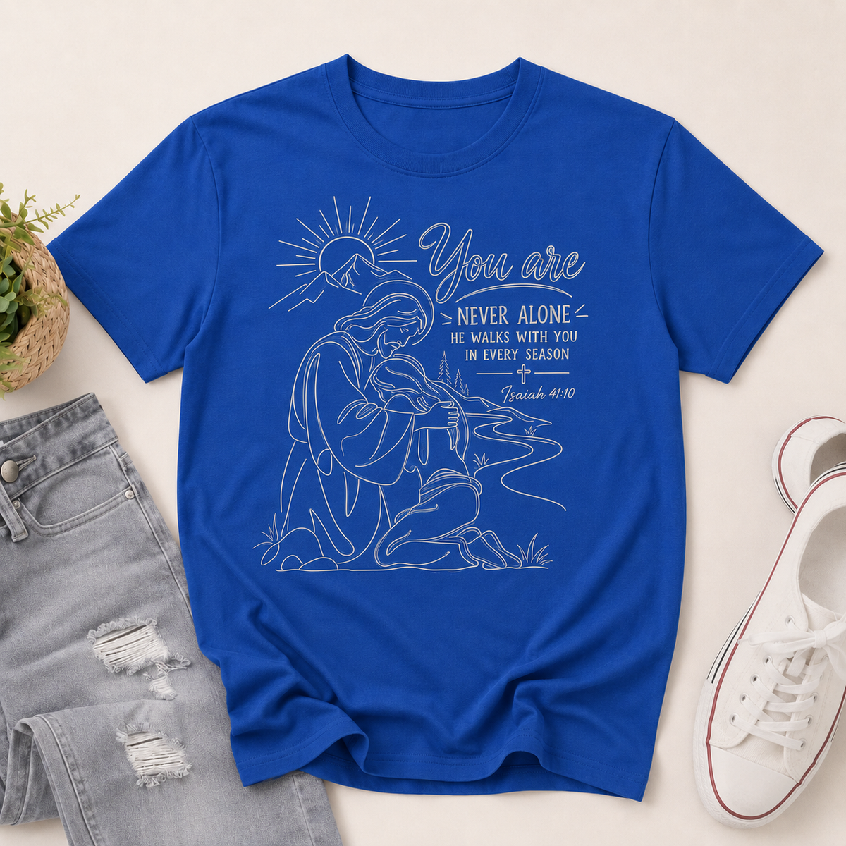 A royal blue short-sleeve graphic T-shirt laid flat on a light beige surface, styled with gray distressed jeans, a small potted plant, and white sneakers. The shirt has a white line-art illustration of a kneeling figure with rays and the text 'You are NEVER ALONE' and a Bible verse.