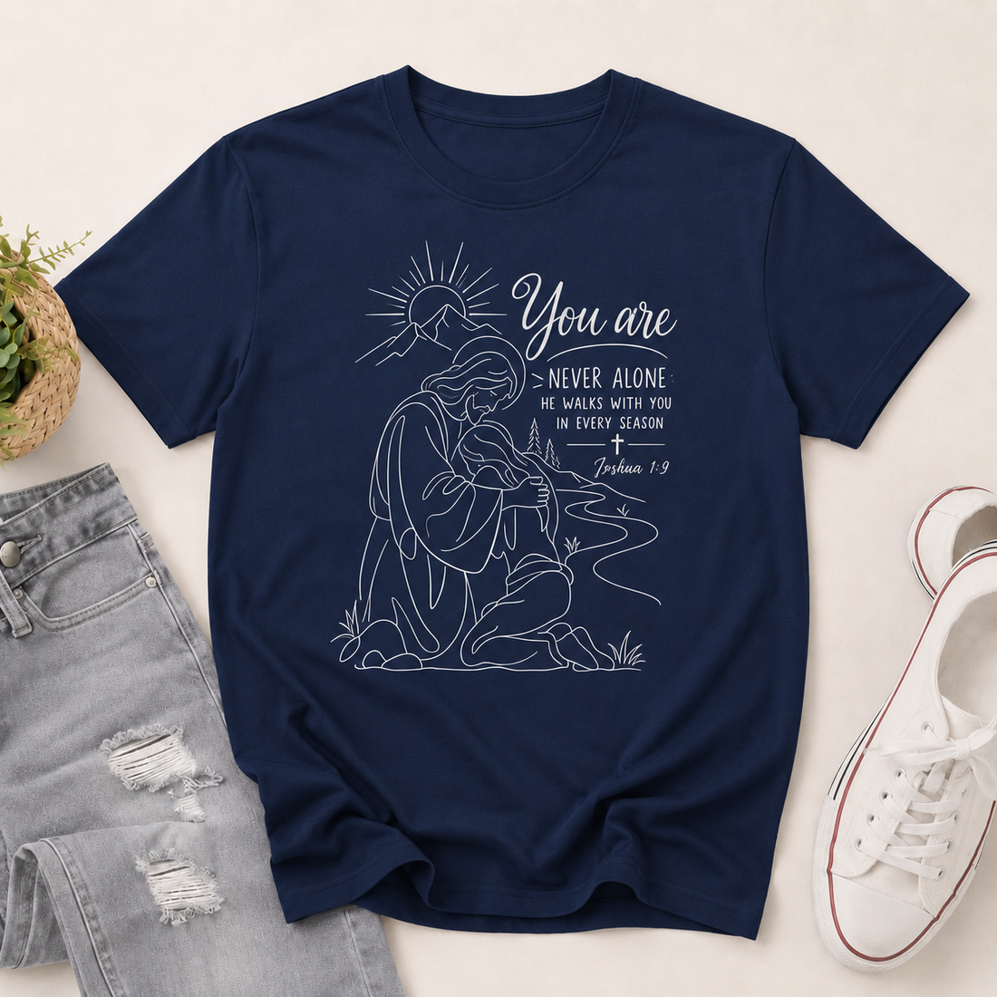 A black short-sleeve graphic T-shirt laid flat on a light beige surface, styled with light-wash distressed jeans, a small potted plant, and a white sneaker. The shirt features a white line-art illustration of a kneeling woman with a sun and the text 'You are NEVER ALONE' and 'He walks with you in every season / Jeremiah 40:15'.
