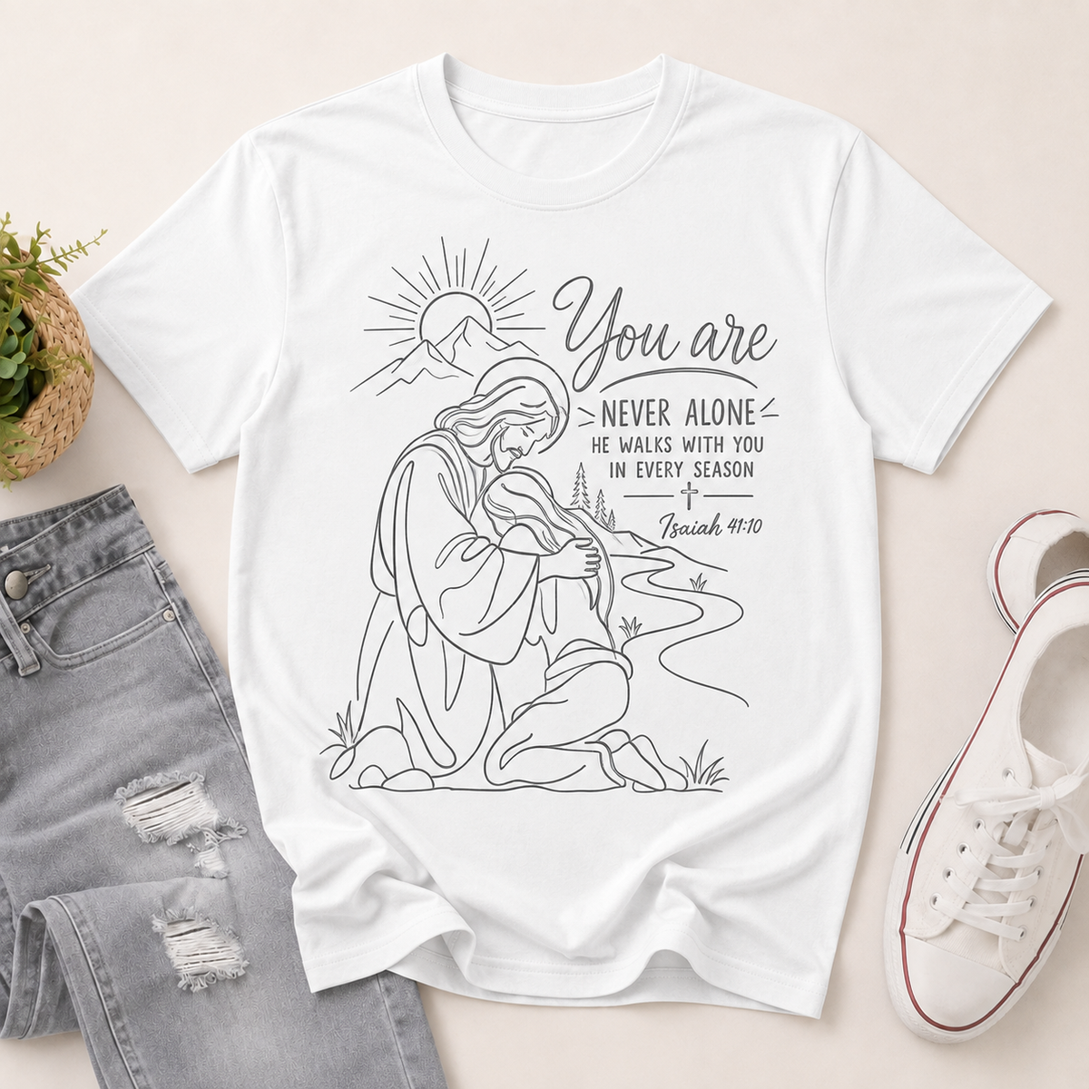 A white graphic T-shirt laid flat on a light beige surface, styled with distressed gray jeans, a small potted plant, and white sneakers. The shirt features a black line drawing of a kneeling man with a sun and river, with the text 'You are NEVER ALONE HE WALKS WITH YOU IN EVERY Season' and 'Isaiah 49:10'.