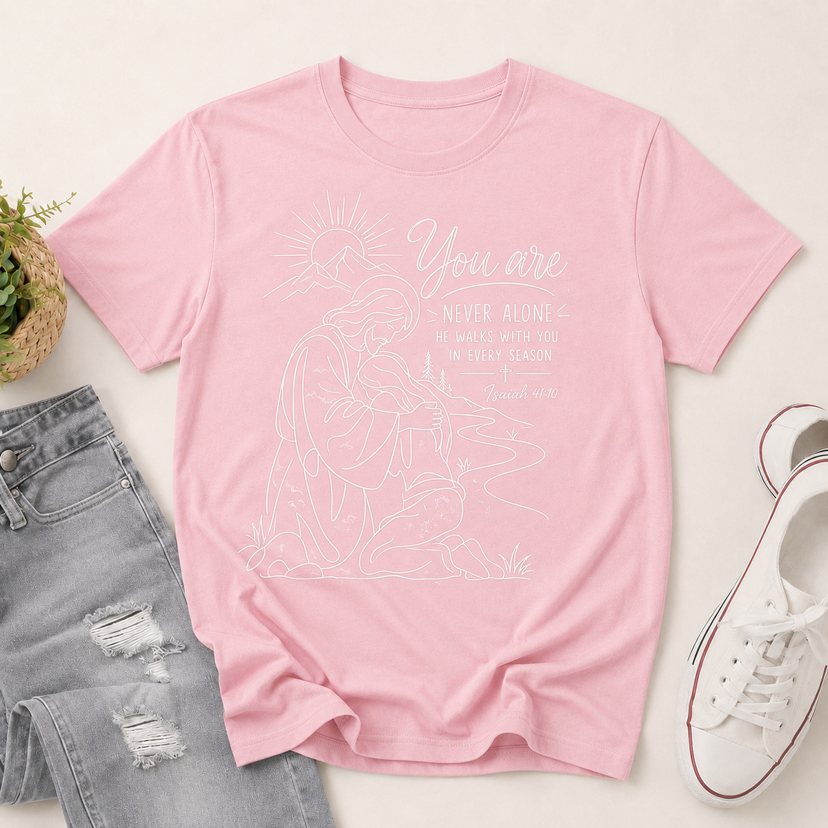 A light pink short-sleeve graphic T-shirt laid flat on a light beige background, styled with distressed gray jeans, a small potted plant, and white sneakers. The shirt features a white line-art illustration of a kneeling woman holding a candle, with the text 'You are NEVER ALONE' and 'IN EVERY SEASON' plus 'Jesus loves you.