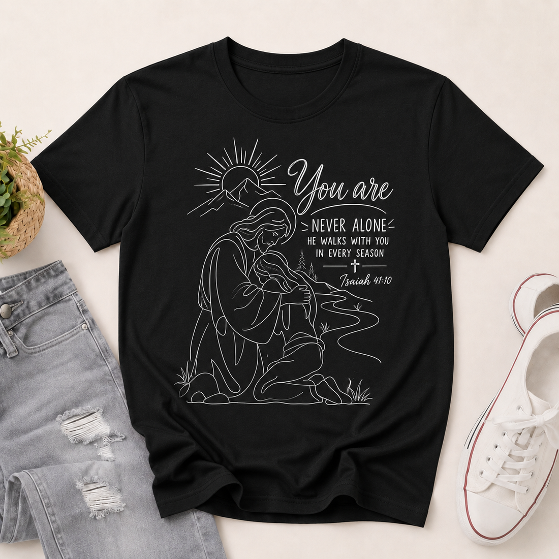 A black short-sleeve graphic T-shirt laid flat on a light beige surface, styled with light-wash distressed jeans, a small potted plant, and a white sneaker. The shirt features a white line-art illustration of a kneeling woman with a sun and the text 'You are NEVER ALONE' and 'He walks with you in every season / Jeremiah 40:15'.