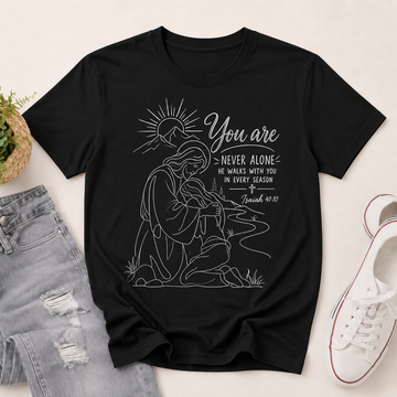 A black short-sleeve graphic T-shirt laid flat on a light beige surface, styled with light-wash distressed jeans, a small potted plant, and a white sneaker. The shirt features a white line-art illustration of a kneeling woman with a sun and the text 'You are NEVER ALONE' and 'He walks with you in every season / Jeremiah 40:15'.